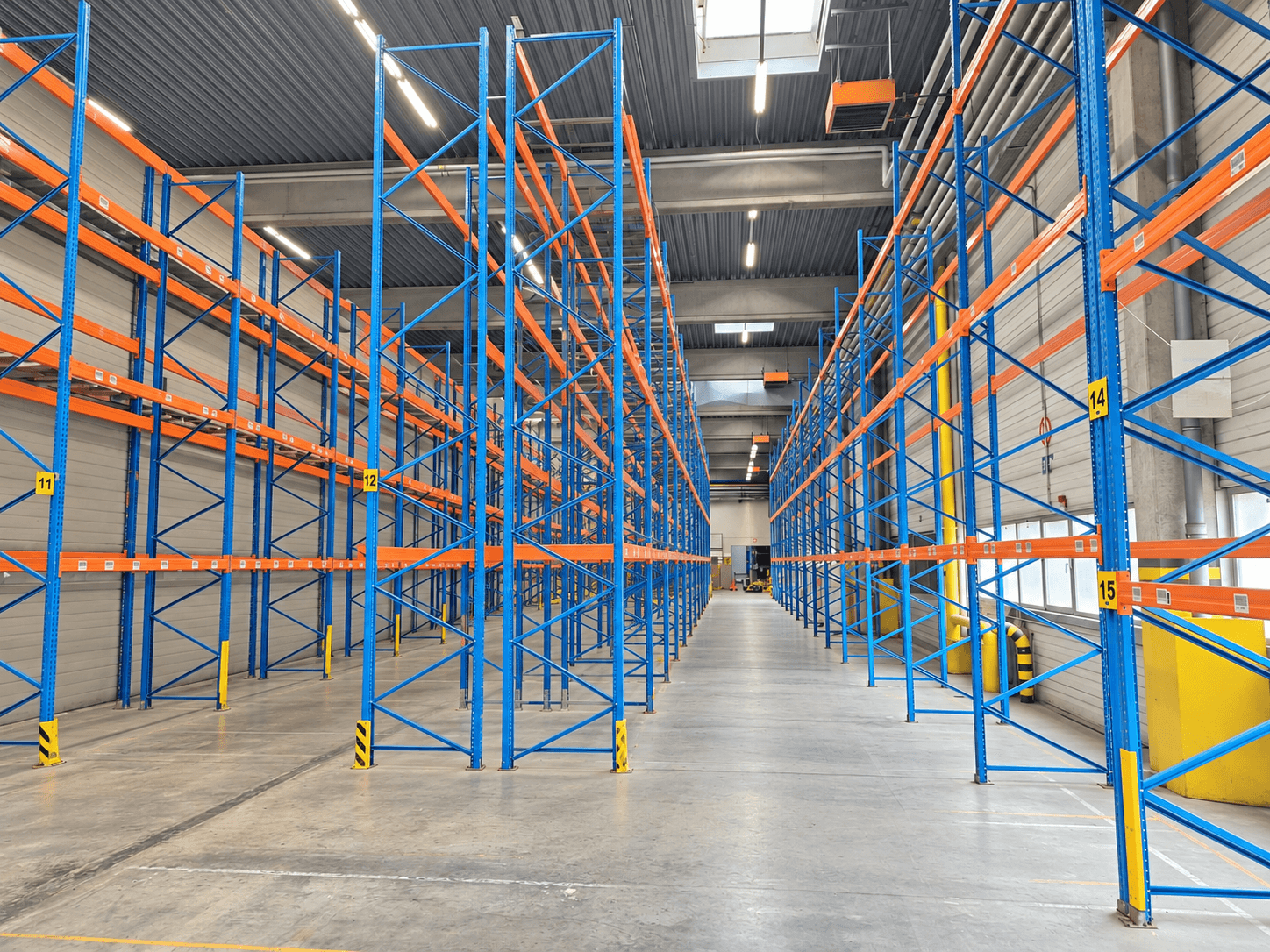 Hohe leere blau-orange Metallregale in einer großen Lagerhalle mit nummerierten Gängen und hellem Industrielicht.