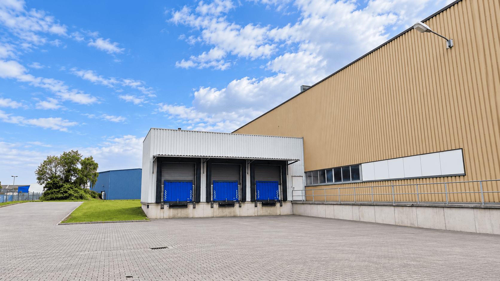 Beigefarbene Lagerhalle mit drei blauen Laderampen auf einem gepflasterten Vorplatz unter blauem Himmel.